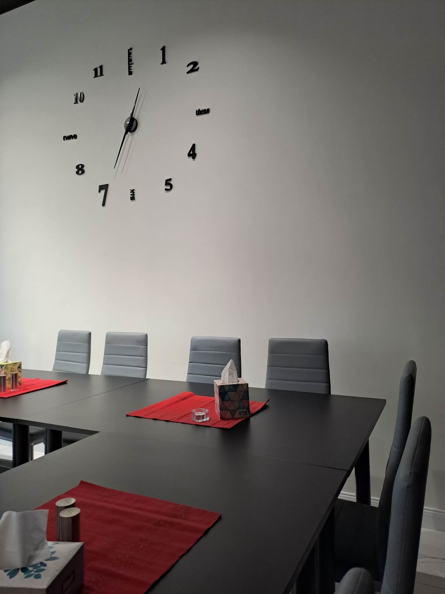Sala konferencyjna z dużym zegarem ściennym, czarnym stołem, szarymi krzesłami i czerwonymi podkładkami pod talerze, pudełko chusteczek higienicznych na stole.