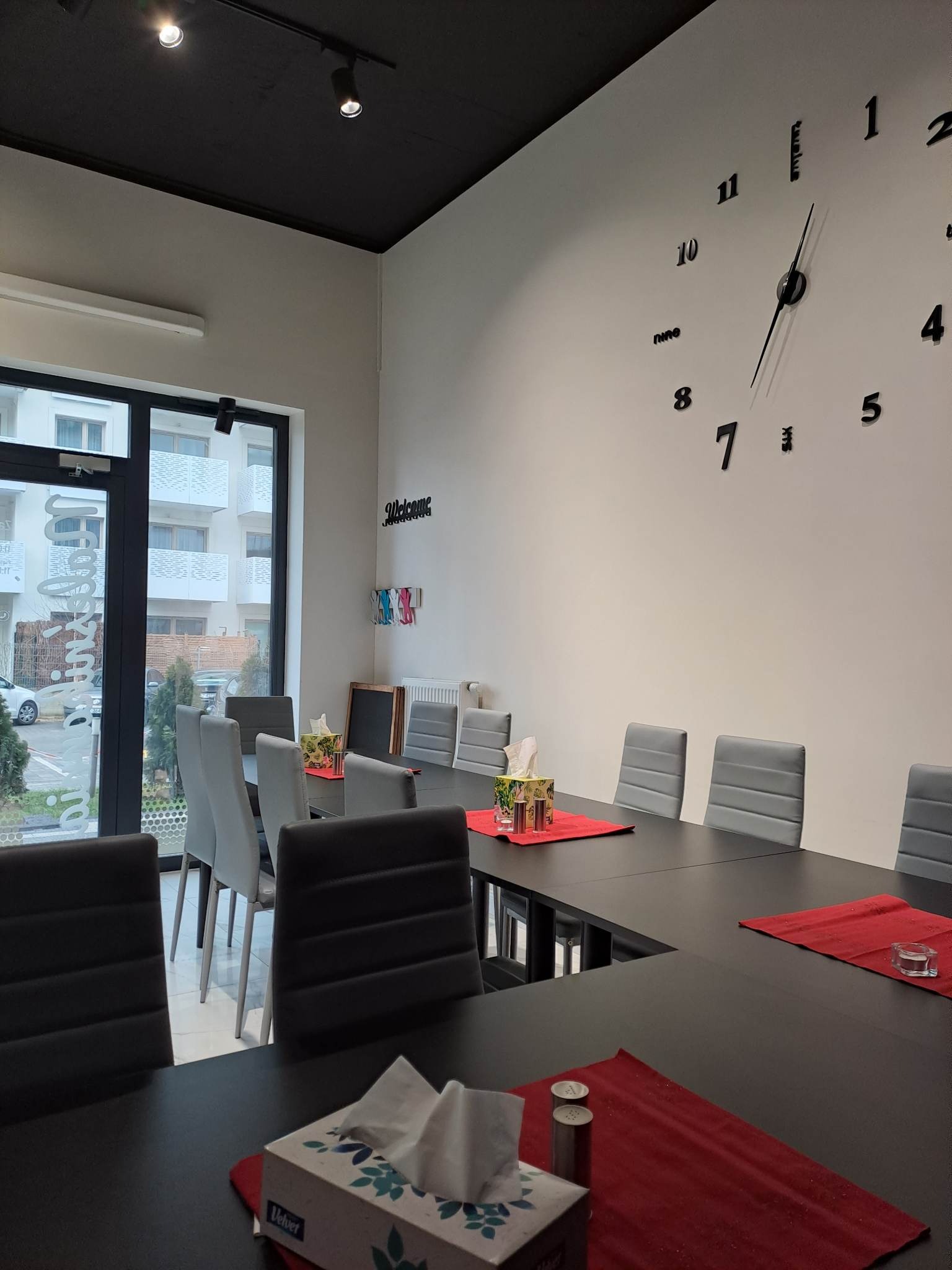 Nowoczesna sala konferencyjna z długim, czarnym stołem, szarymi krzesłami, dekoracyjnym zegarem na ścianie i pudełkami chusteczek na czerwonych podkładkach.