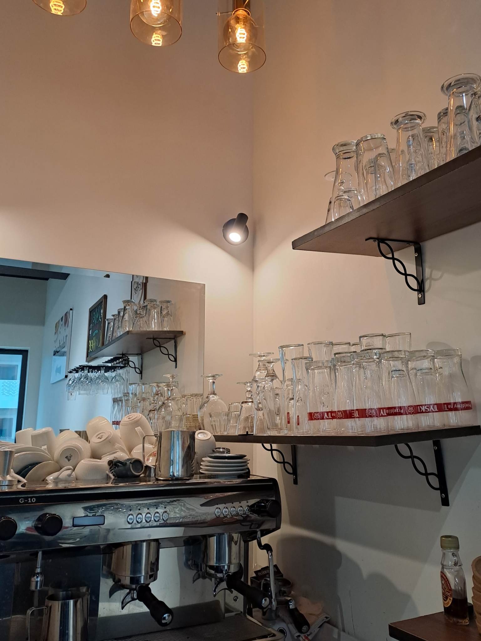 Bar z profesjonalnym ekspresem do kawy, stertą białych filiżanek i talerzyków, oraz półkami z czystymi szklankami i kieliszkami do wina, oświetlony ciepłym światłem żarówek.