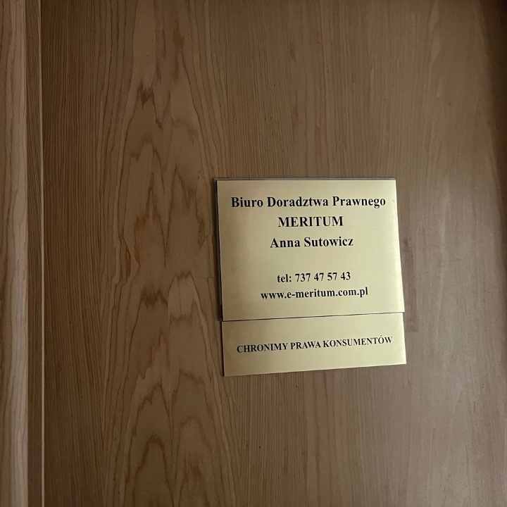 Tabliczka informacyjna na drewnianych drzwiach z napisem 'Biuro Doradztwa Prawnego MERITUM Anna Sutowicz' oraz danymi kontaktowymi i hasłem 'CHRONIMY PRAWA KONSUMENTÓW'.