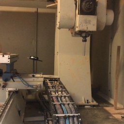 Global Mechatronics Service - Wnętrze warsztatu z 5-osiowym centrum obróbczym FANUM, w trakcie kalibracji osi za pomocą specjalistycznego przyrządu i czujnika zegarowego. Widoczny stos obrobionych elementów drewnianych.