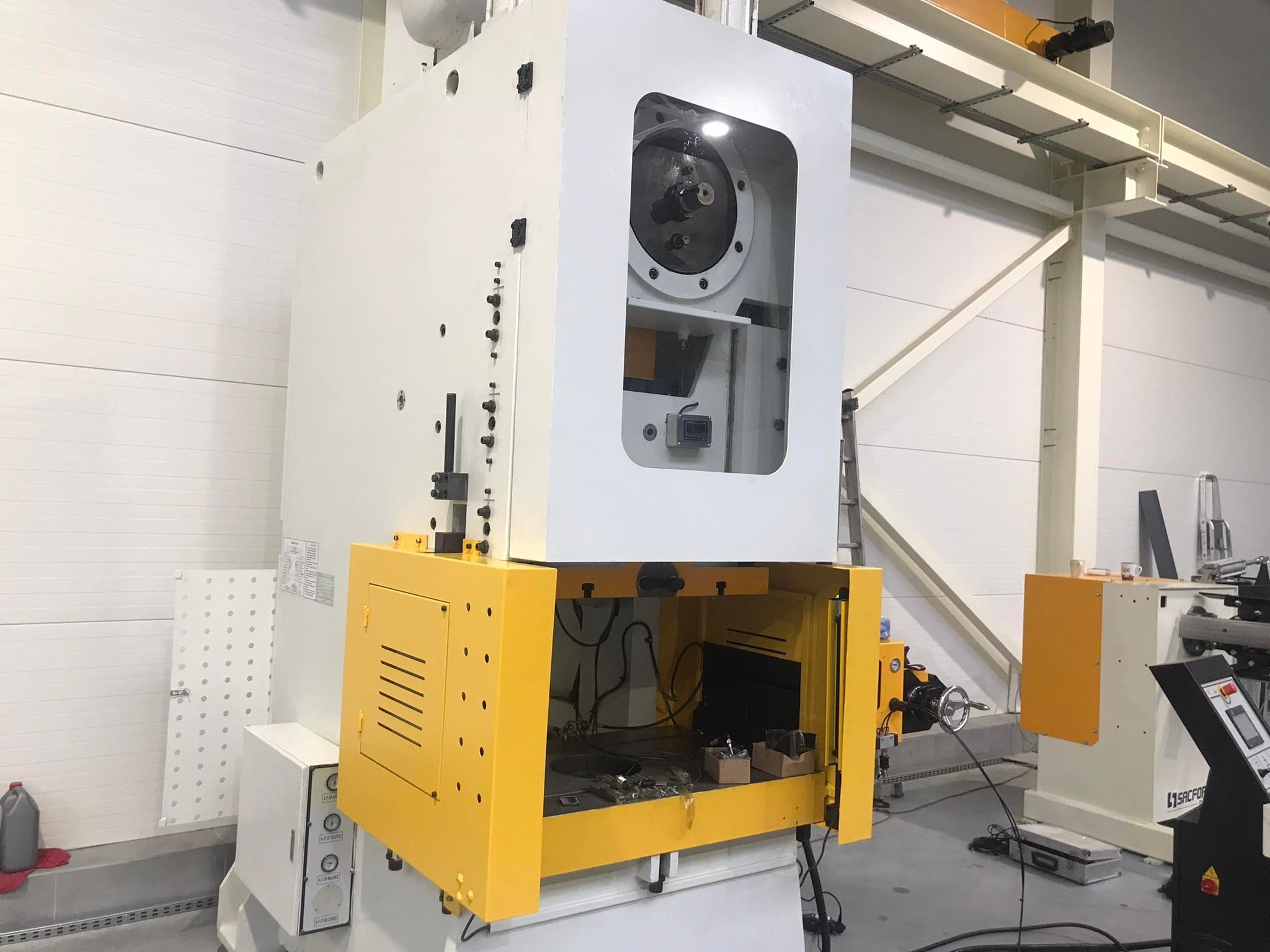 Duża, biało-żółta prasa mechaniczna w hali produkcyjnej z widocznymi elementami wewnętrznymi i narzędziami, oświetlona światłem dziennym.