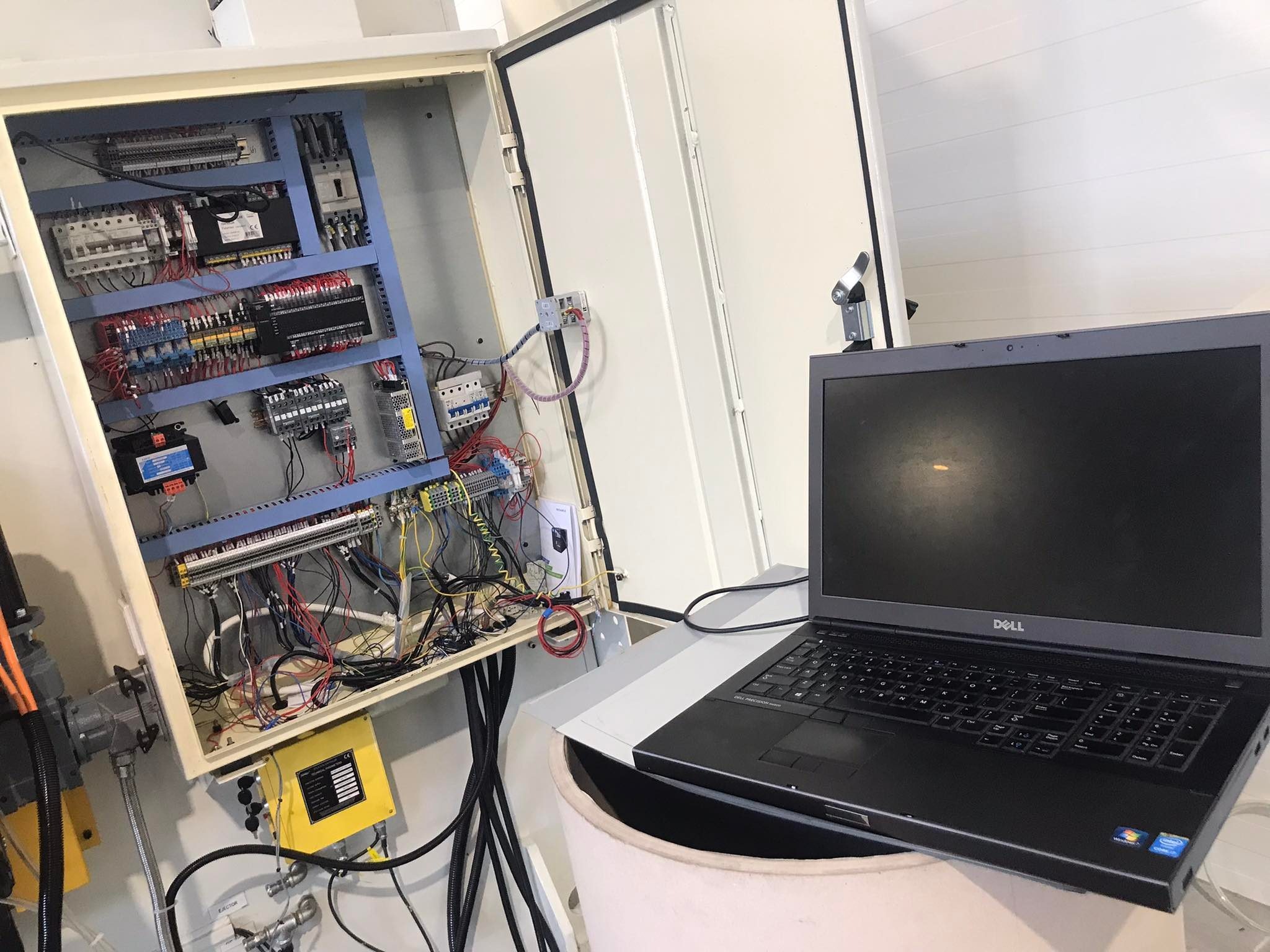Otwarta szafa sterownicza z widocznymi elementami elektrycznymi i przewodami, obok laptop Dell Precision, prawdopodobnie używany do diagnostyki lub programowania.