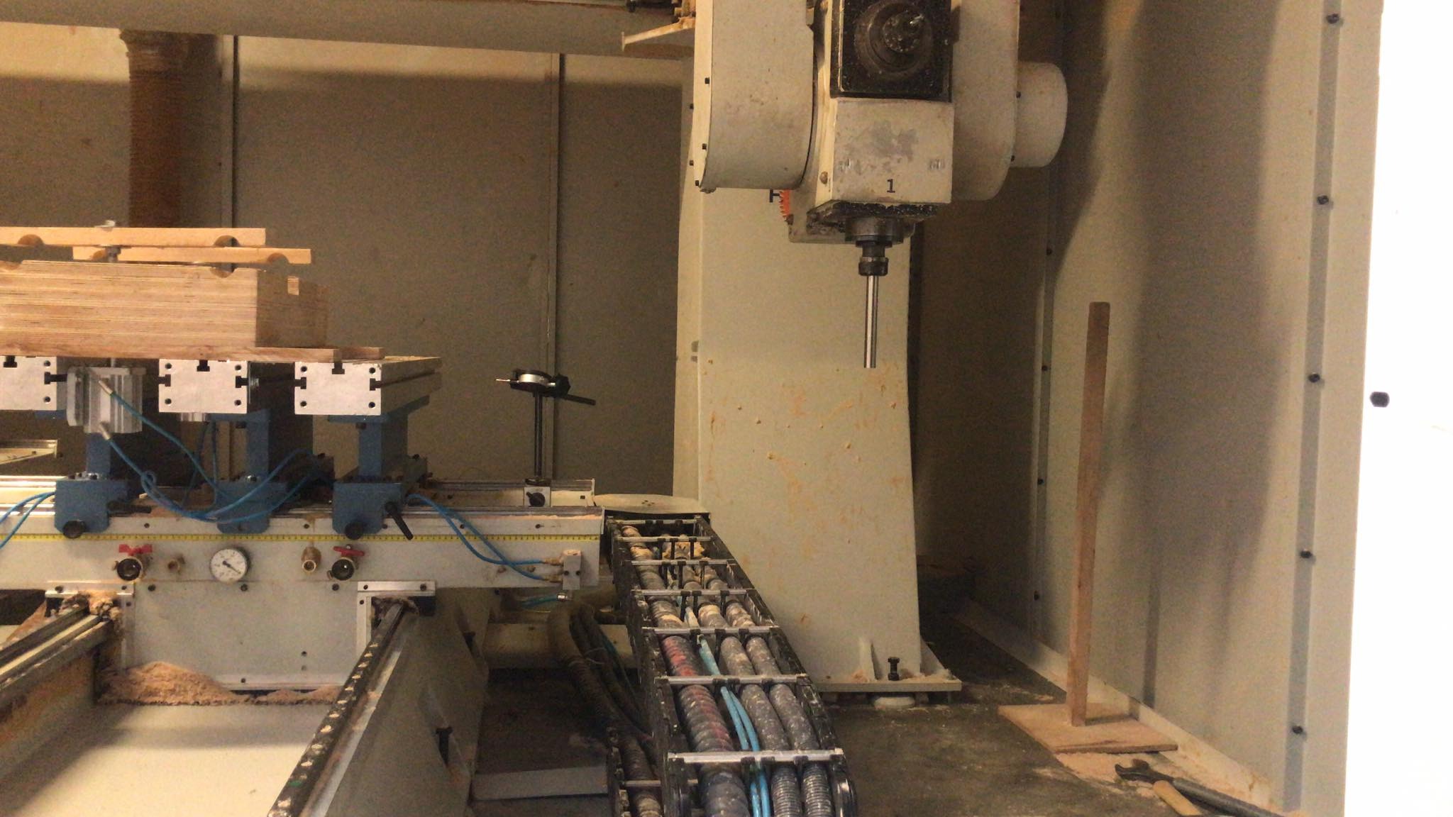 Wnętrze warsztatu z 5-osiowym centrum obróbczym FANUM, w trakcie kalibracji osi za pomocą specjalistycznego przyrządu i czujnika zegarowego. Widoczny stos obrobionych elementów drewnianych.