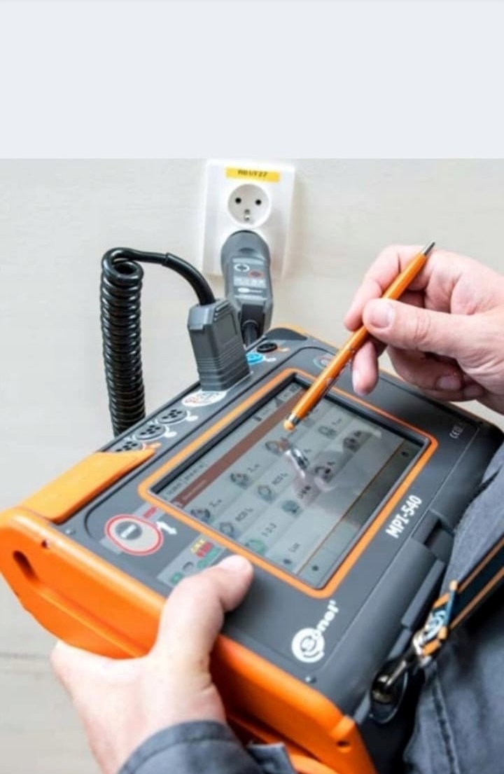 Elektryk używa miernika MPI-540 do pomiaru parametrów gniazdka elektrycznego, widoczna wtyczka adaptera w gniazdku i spiralny kabel pomiarowy.