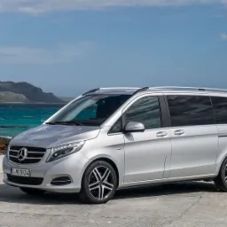 Srebrny Mercedes-Benz V-Class zaparkowany na betonowej powierzchni z widokiem na turkusowe morze i górzysty krajobraz w tle.