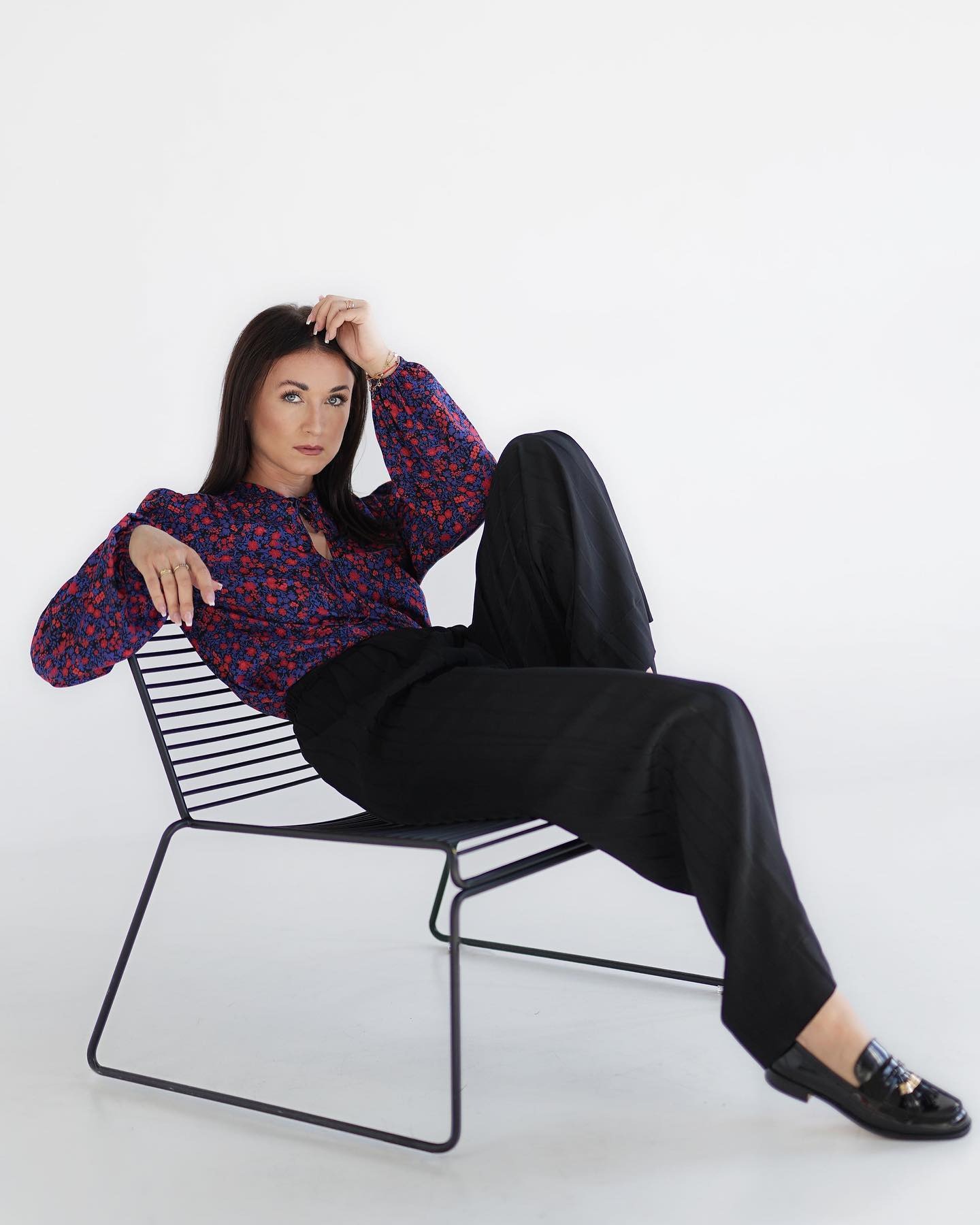 Kobieta siedząca na metalowym krześle, ubrana w kwiecistą bluzkę z długim rękawem i szerokie czarne spodnie, prezentuje stylizację z lakierowanymi mokasynami z frędzlami.