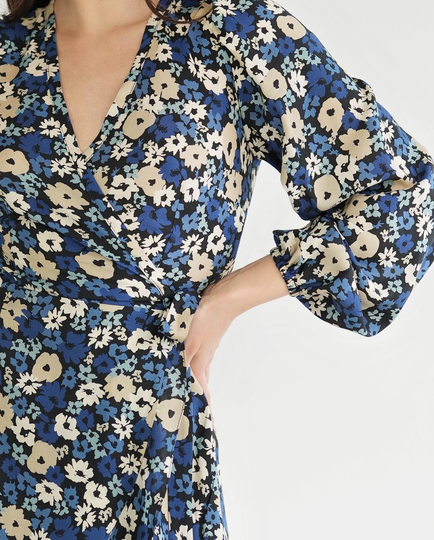 Kobieta w kwiecistej sukience z kopertowym dekoltem i bufiastymi rękawami, pozująca na białym tle, ręka na biodrze.