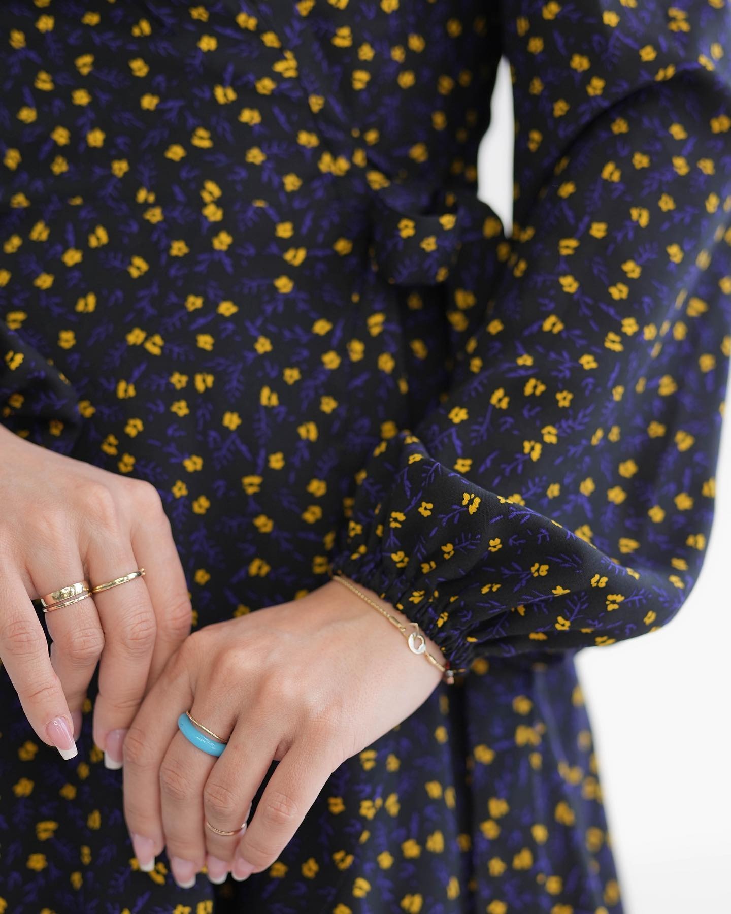 Fragment sukienki w drobne żółte kwiatki na granatowym tle, z widocznymi dłońmi modelki ozdobionymi złotymi pierścionkami i turkusowym oczkiem.