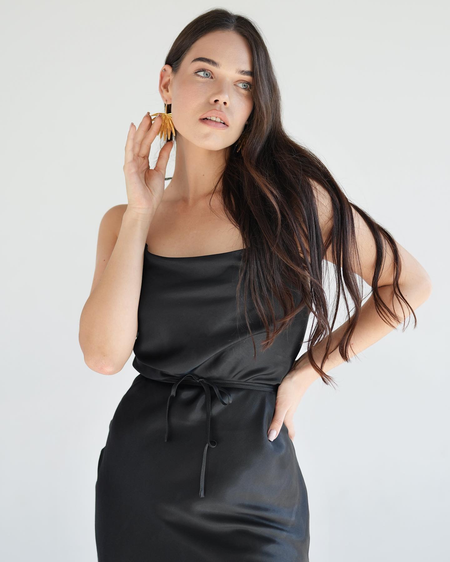 Brunetka w czarnej, satynowej sukience na ramiączkach, z ręką przy twarzy, eksponująca złote kolczyki w kształcie liści.