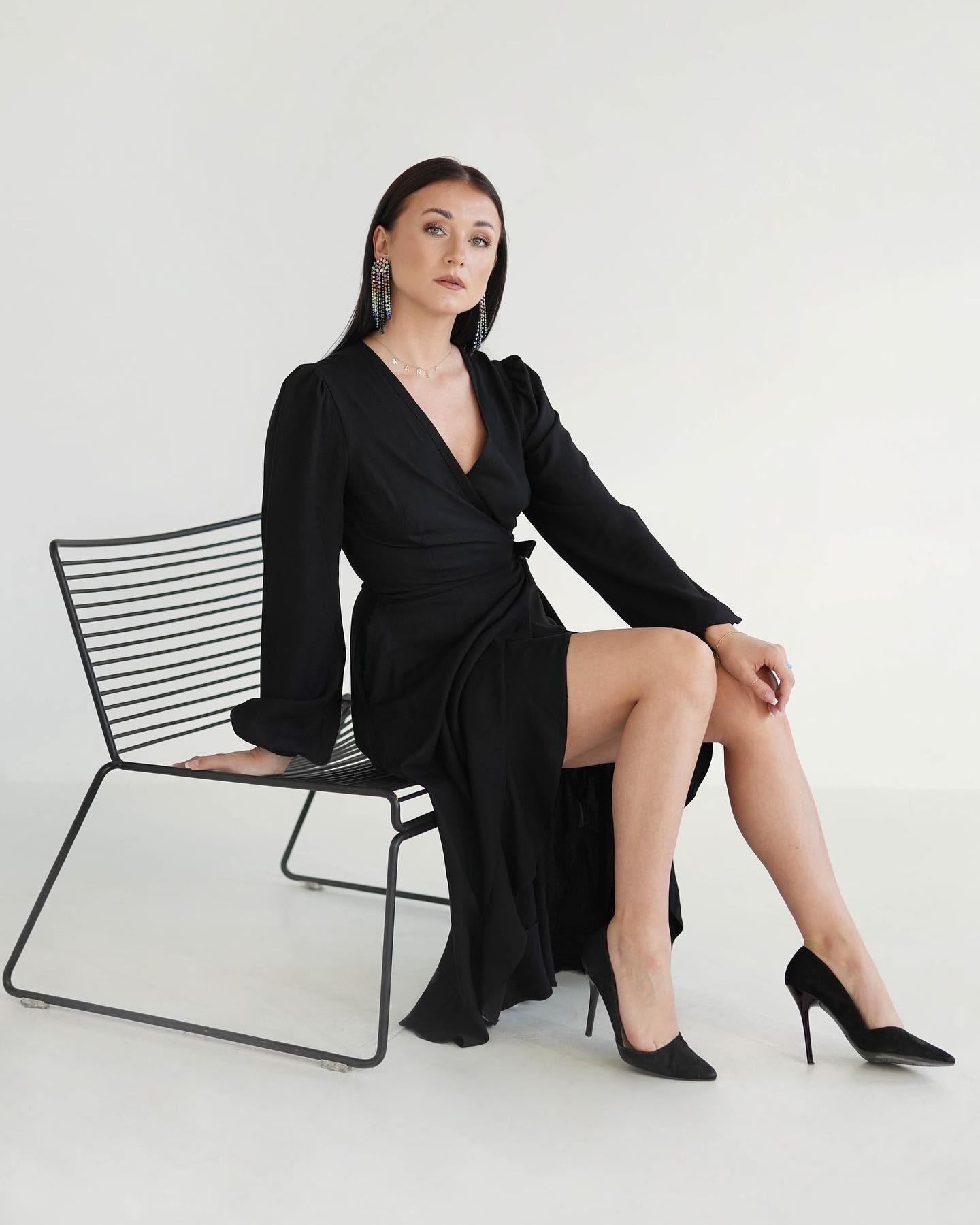 Kobieta siedząca na metalowym krześle w czarnej sukience z kopertowym dekoltem, długimi rękawami i czarnych szpilkach, z wiszącymi kolczykami z kolorowych koralików i naszyjnikiem z imieniem.