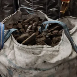 Worek typu big bag wypełniony brykietem o nieregularnych kształtach, widoczne niebieskie uchwyty, w tle ciemne worki z opałem.