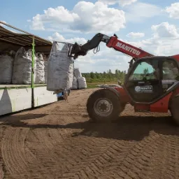 Czerwony wózek widłowy Manitou podnosi duży, biały worek z materiałem na przyczepę ciężarówki; w tle widoczne inne worki i zielone drzewa.