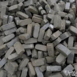 Stos szarych, prostokątnych brykietów węglowych ułożonych chaotycznie, widoczne nierówności i pęknięcia na powierzchni materiału.