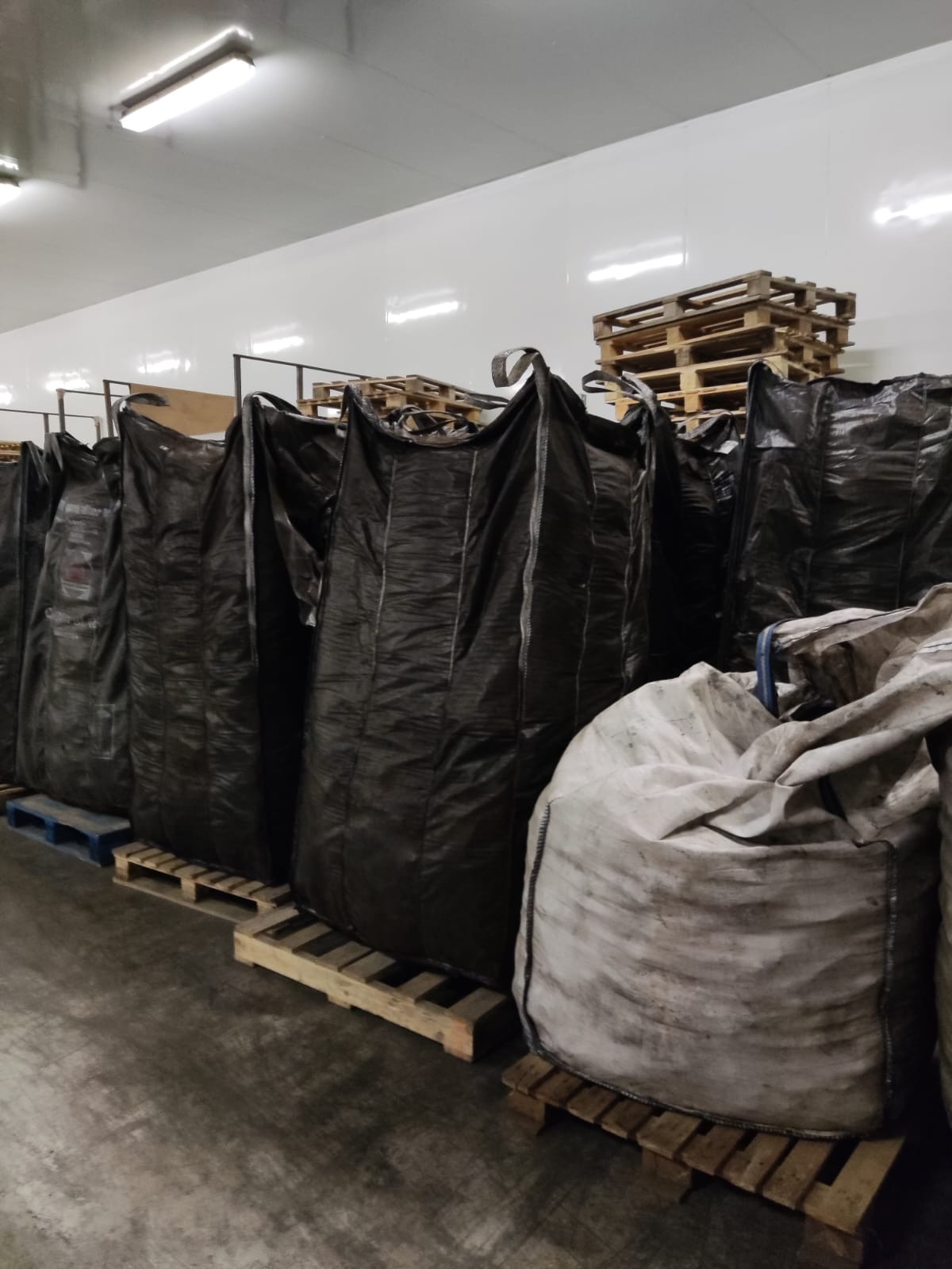 Rząd worków typu big bag, częściowo czarnych, częściowo szarych, na paletach w magazynie z białymi ścianami i oświetleniem jarzeniowym.