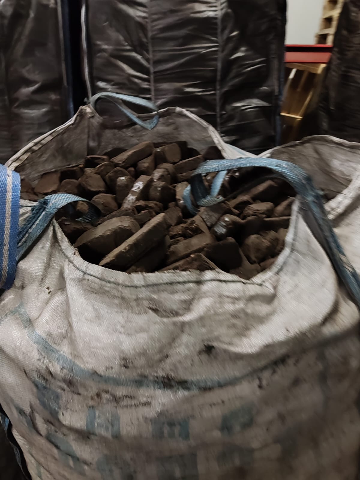 Worek typu big bag wypełniony brykietem o nieregularnych kształtach, widoczne niebieskie uchwyty, w tle ciemne worki z opałem.