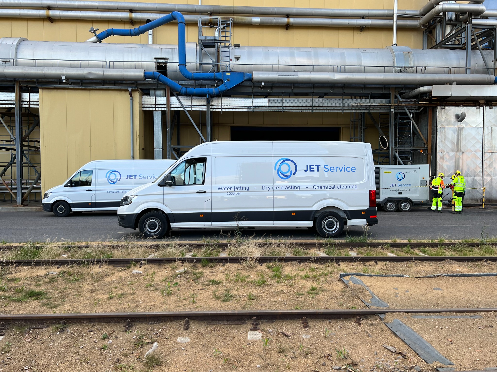 Dwa białe busy firmy Jet Service z logiem i opisem usług (water jetting, dry ice blasting, chemical cleaning) zaparkowane przed przemysłowym budynkiem z rurami, w tle generator i dwóch pracowników...