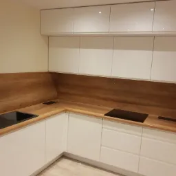 Nowoczesna kuchnia w kształcie litery L z białymi, lakierowanymi frontami szafek i blatem z drewnopodobnym wzorem, zintegrowany z czarnym zlewem i płytą indukcyjną.