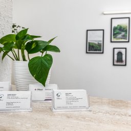 Kancelaria Gospodarcza EFFEKTI sp. z o.o. - Wizytówki firmowe w przezroczystych stojakach na marmurowym blacie, z rośliną w doniczce w tle i ramkami ze zdjęciami na ścianie.