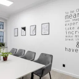 Jasne, minimalistyczne biuro w Poznaniu z białym stołem konferencyjnym, szarymi krzesłami, roślinami doniczkowymi i ścianą ozdobioną mapami miast oraz inspirującym napisem 'to be CREATIVE means...'.