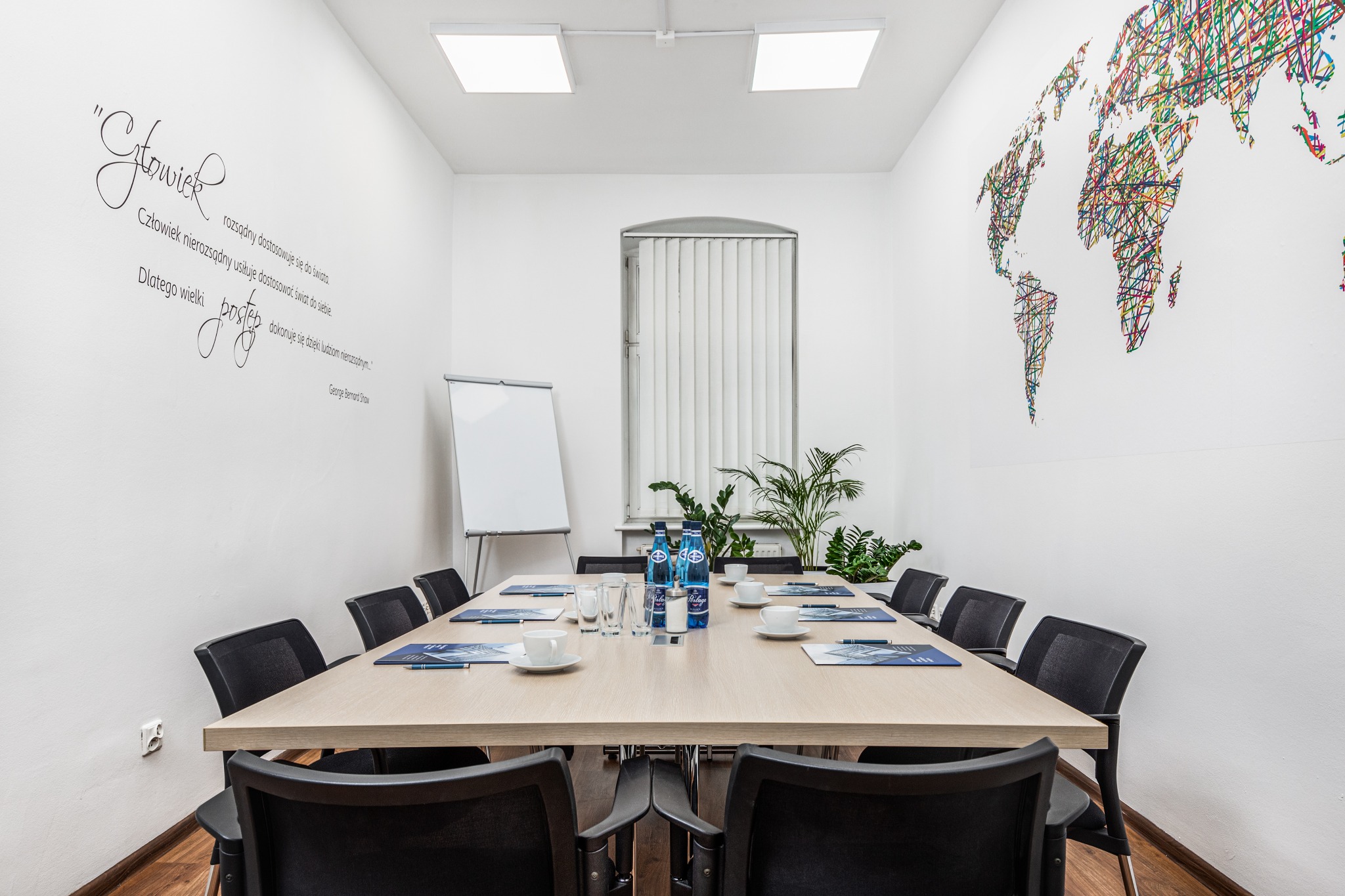 Sala konferencyjna z dużym stołem, krzesłami, wodą, filiżankami i tablicą. Na ścianie cytat i kolorowa mapa świata.