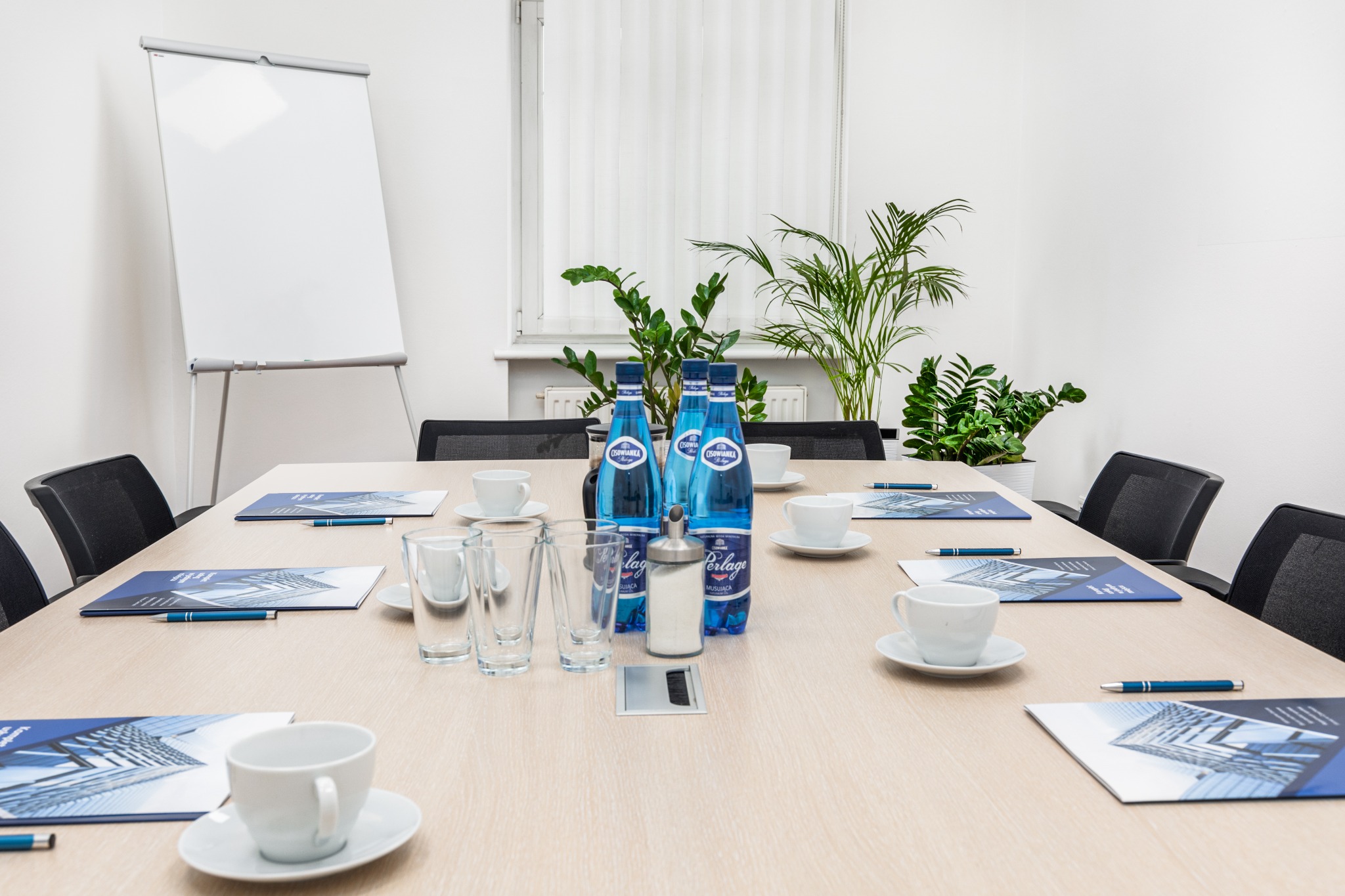 Sala konferencyjna z drewnianym stołem, na którym leżą materiały firmowe, długopisy, szklanki, filiżanki, cukiernica i woda w butelkach, a w tle widoczne są rośliny doniczkowe i tablica...