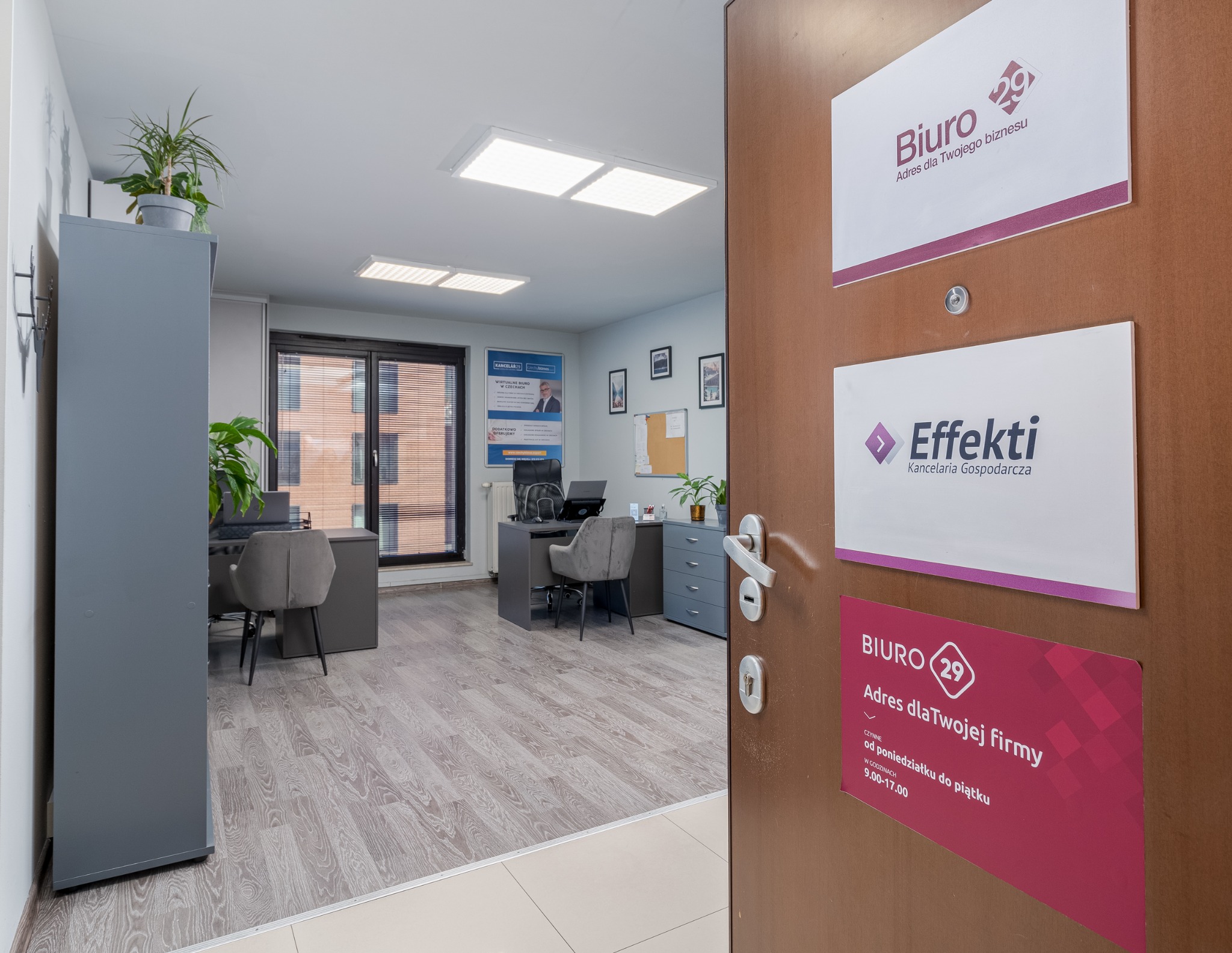 Wnętrze biura z szarymi meblami i roślinami doniczkowymi, widoczne przez uchylone drzwi z tabliczkami informacyjnymi 'Biuro 29' i 'Effekti Kancelaria Gospodarcza'.