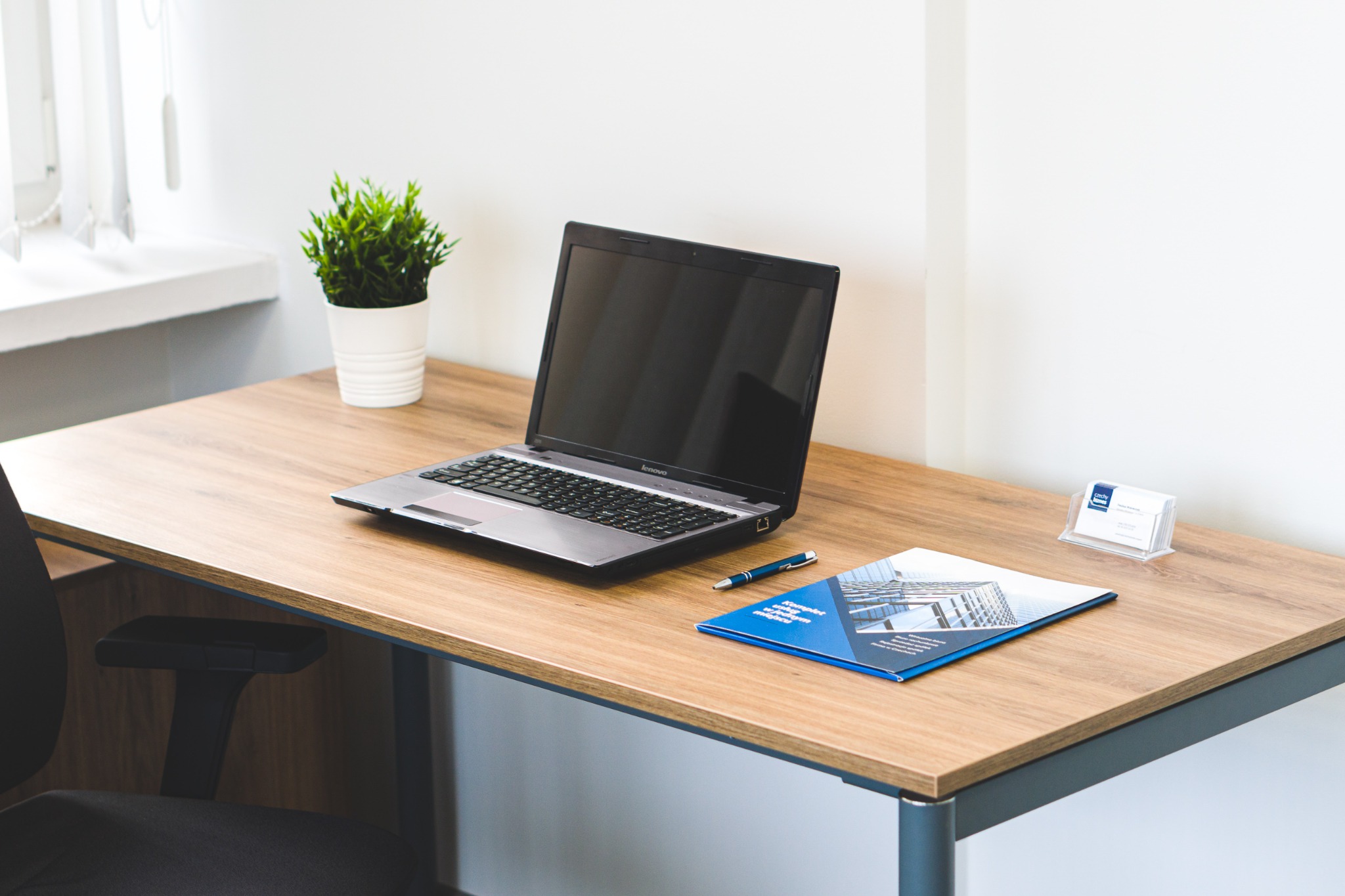 Biurko z laptopem, ulotką reklamową z motywem biurowca, długopisem i wizytownikiem na tle białej ściany i doniczki z rośliną.