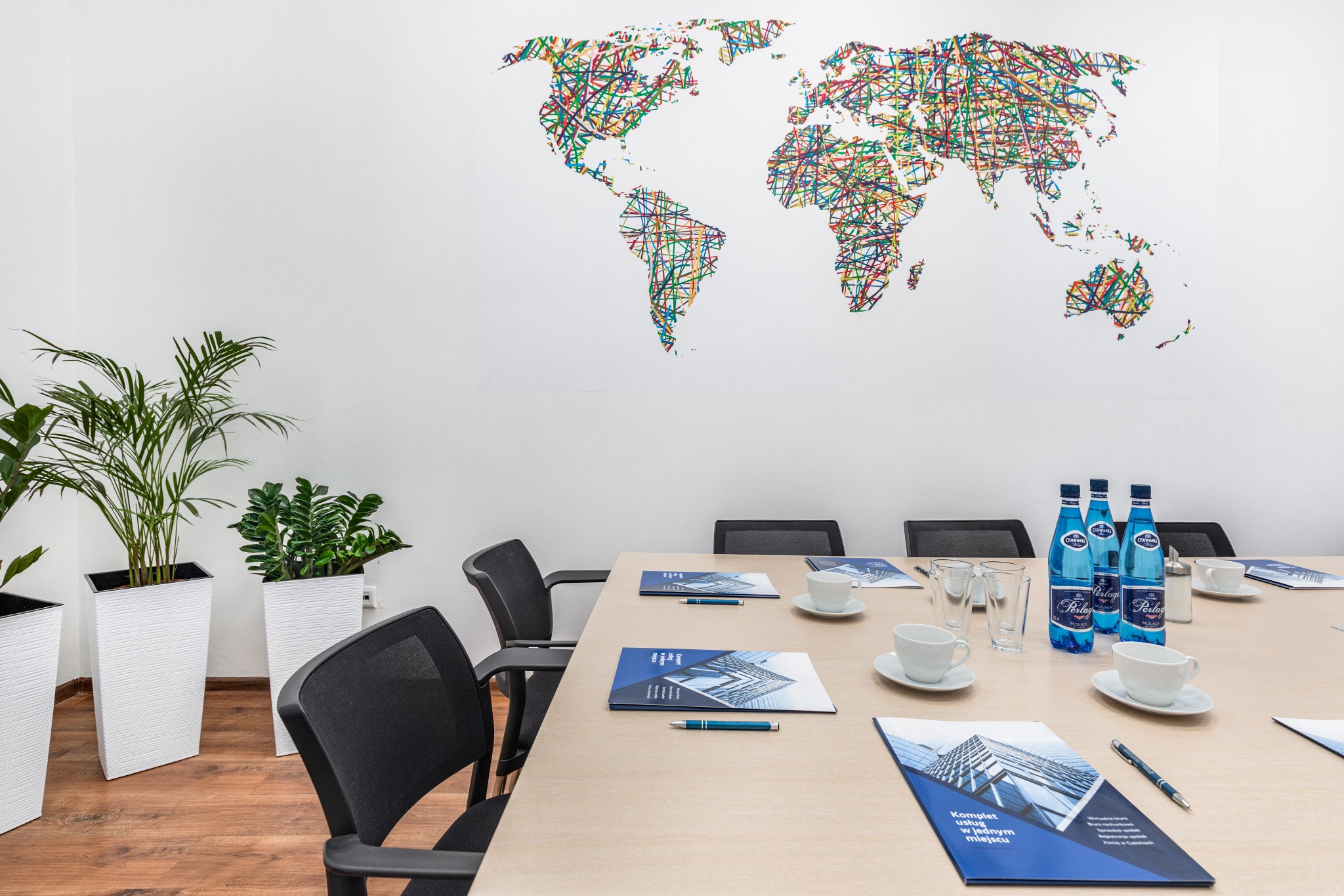 Sala konferencyjna z długim stołem, krzesłami, roślinami doniczkowymi i mapą świata z kolorowych linii na ścianie, przygotowana na spotkanie biznesowe z broszurami, długopisami, wodą i filiżankami.