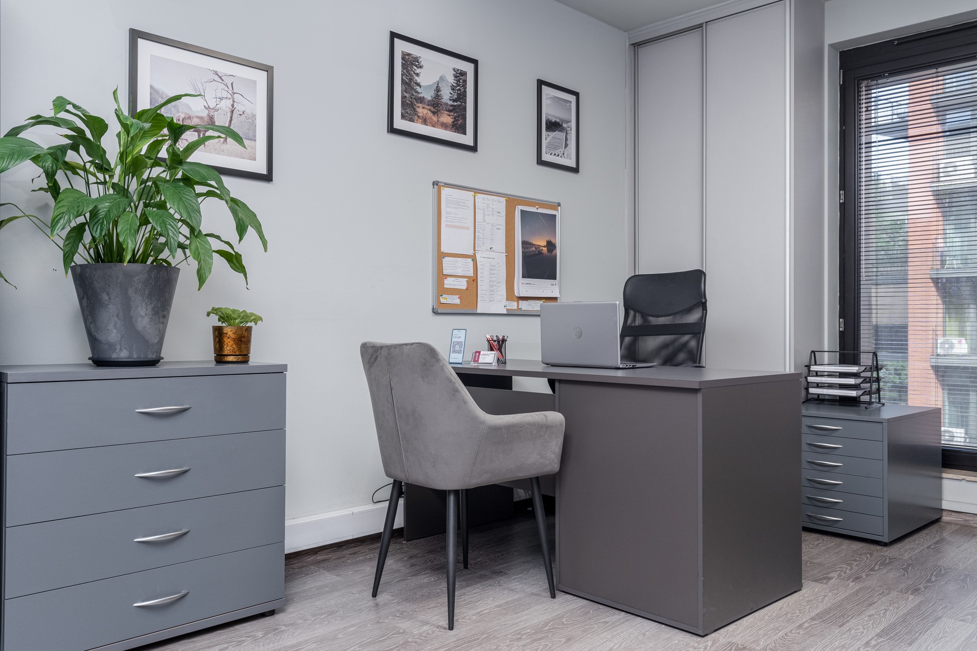 Nowoczesne biuro: Szary biurko z laptopem, krzesłem i tablicą korkową, uzupełnione roślinami doniczkowymi i minimalistycznymi obrazami.