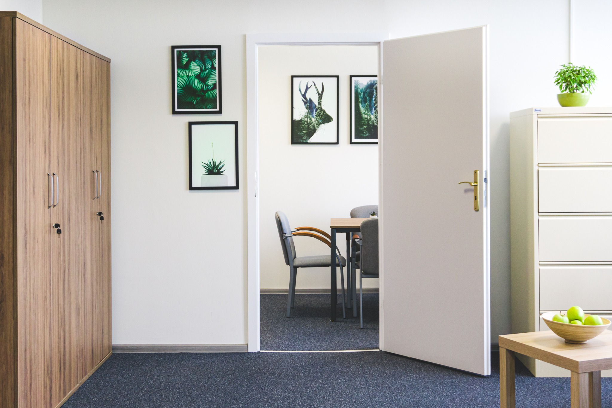 Jasne biuro z otwartymi drzwiami do sali konferencyjnej, na ścianach grafiki z motywami roślinnymi i zwierzęcymi, drewniana szafa po lewej, a po prawej szafka z zieloną rośliną i stolik z owocami.