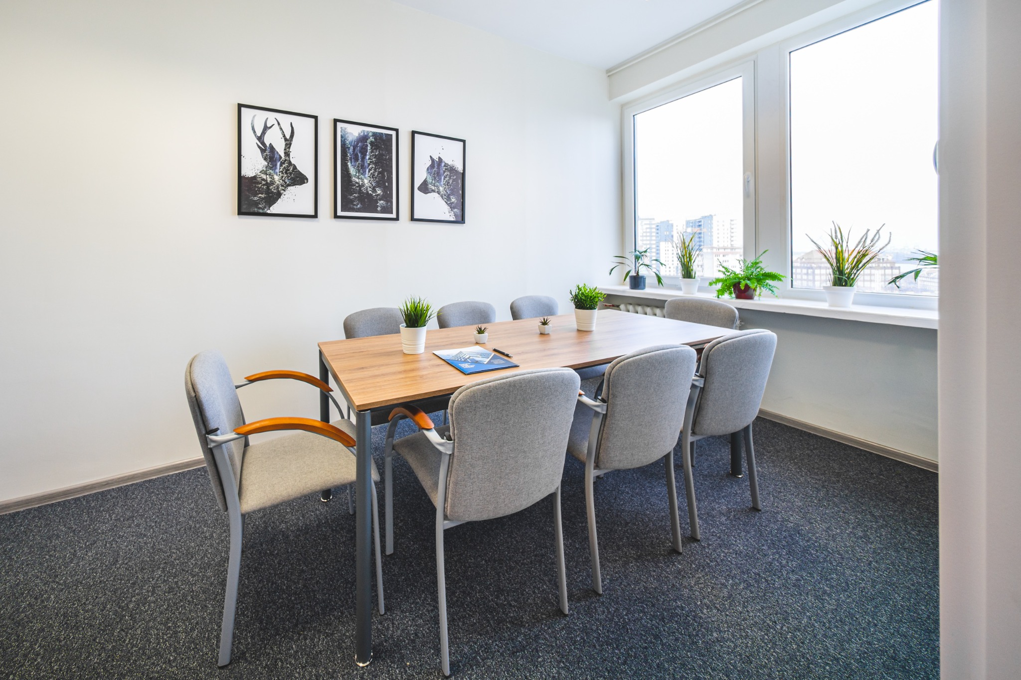 Jasna sala konferencyjna z drewnianym stołem, szarymi krzesłami, trzema obrazami zwierząt na ścianie i roślinami w doniczkach na parapecie okiennym z widokiem na miasto.