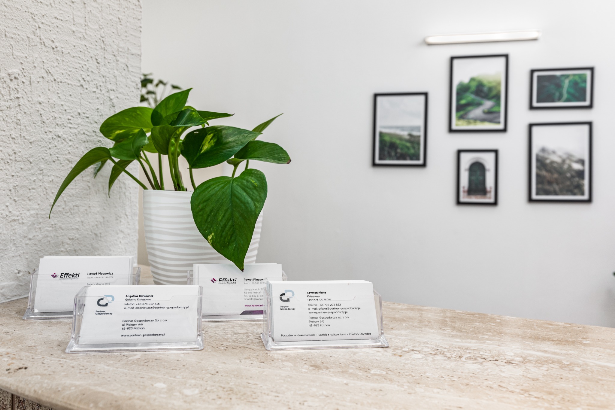 Wizytówki firmowe w przezroczystych stojakach na marmurowym blacie, z rośliną w doniczce w tle i ramkami ze zdjęciami na ścianie.