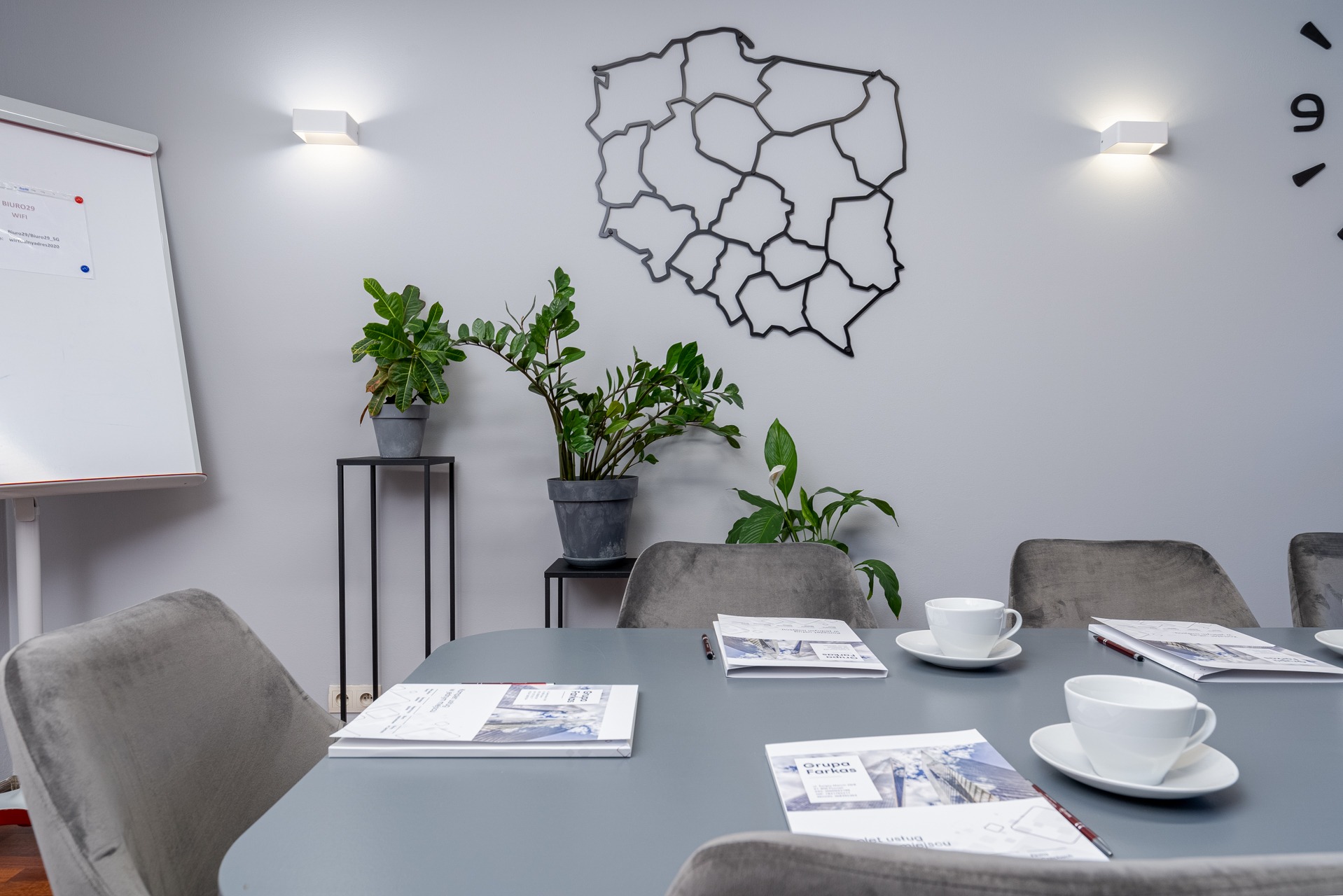 Sala konferencyjna z szarym stołem, welurowymi krzesłami, filiżankami kawy, ulotkami firmowymi, tablicą suchościeralną, roślinami doniczkowymi oraz dekoracją ścienną w kształcie mapy Polski.