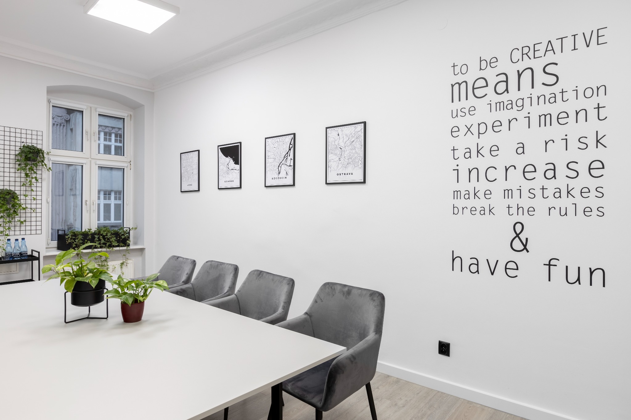 Jasne, minimalistyczne biuro w Poznaniu z białym stołem konferencyjnym, szarymi krzesłami, roślinami doniczkowymi i ścianą ozdobioną mapami miast oraz inspirującym napisem 'to be CREATIVE means...'.