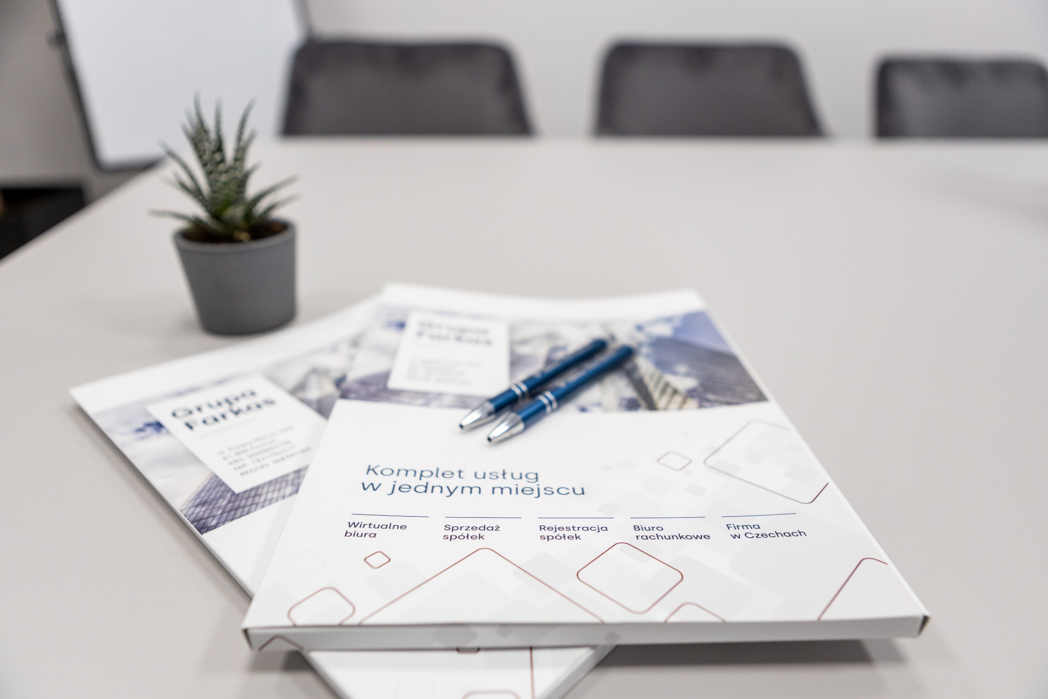 Dwa niebieskie długopisy leżą na broszurze z napisem 'Komplet usług w jednym miejscu', obok stoi mała roślina doniczkowa, w tle niewyraźne krzesła.