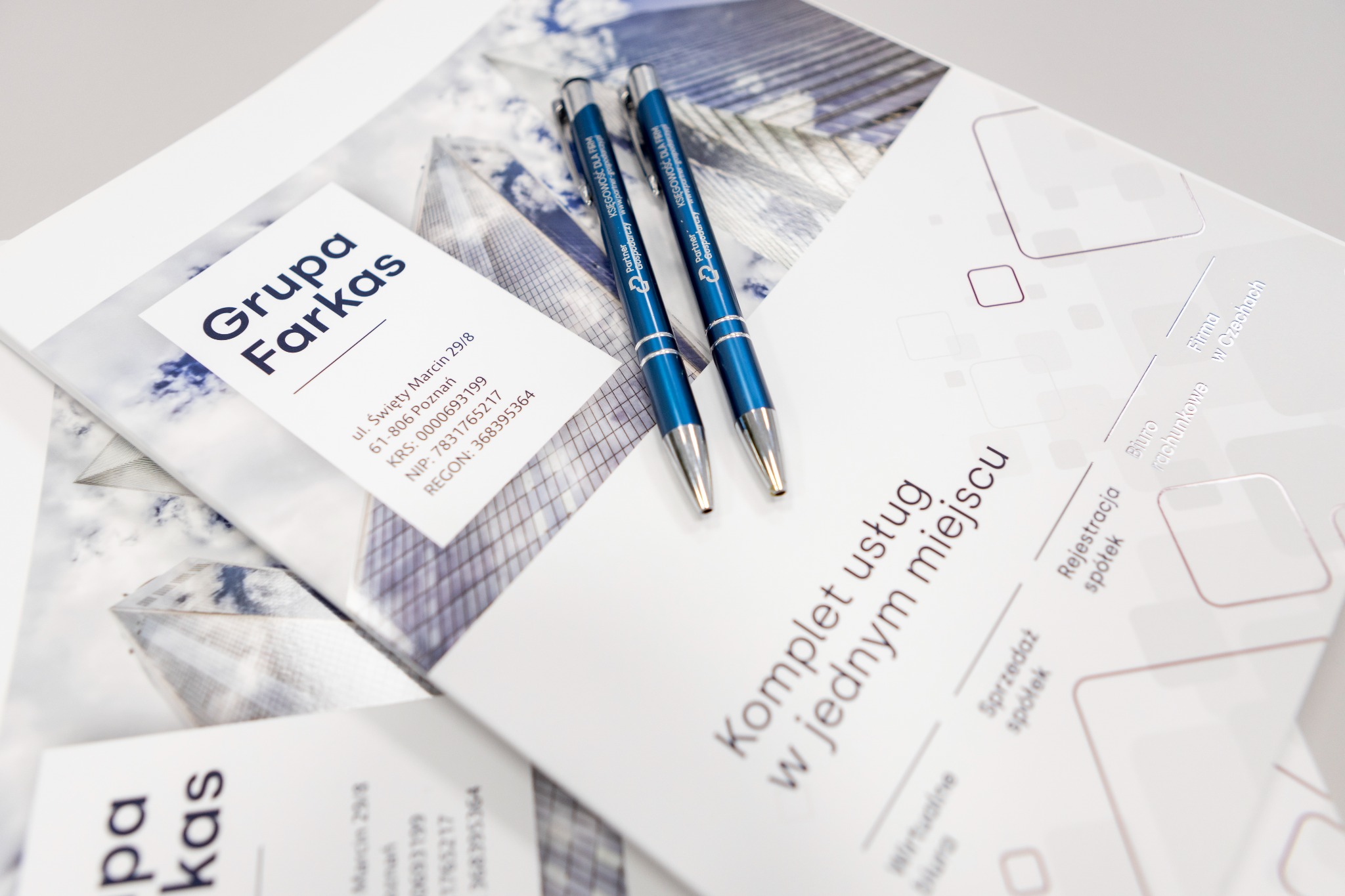 Dwa niebieskie długopisy z logo firmy Partner Gospodarczy leżą na materiałach reklamowych Grupy Farkas oferującej kompleks usług w jednym miejscu, w tym rejestrację i sprzedaż spółek oraz biuro...