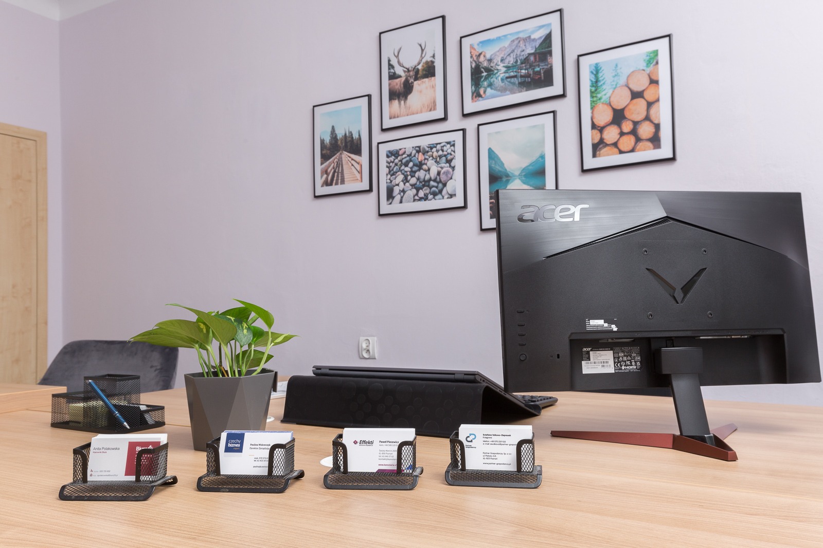 Biurko z monitorem Acer, tabletem, rośliną doniczkową i wizytownikami z wizytówkami na tle ściany z sześcioma obrazkami w czarnych ramkach.