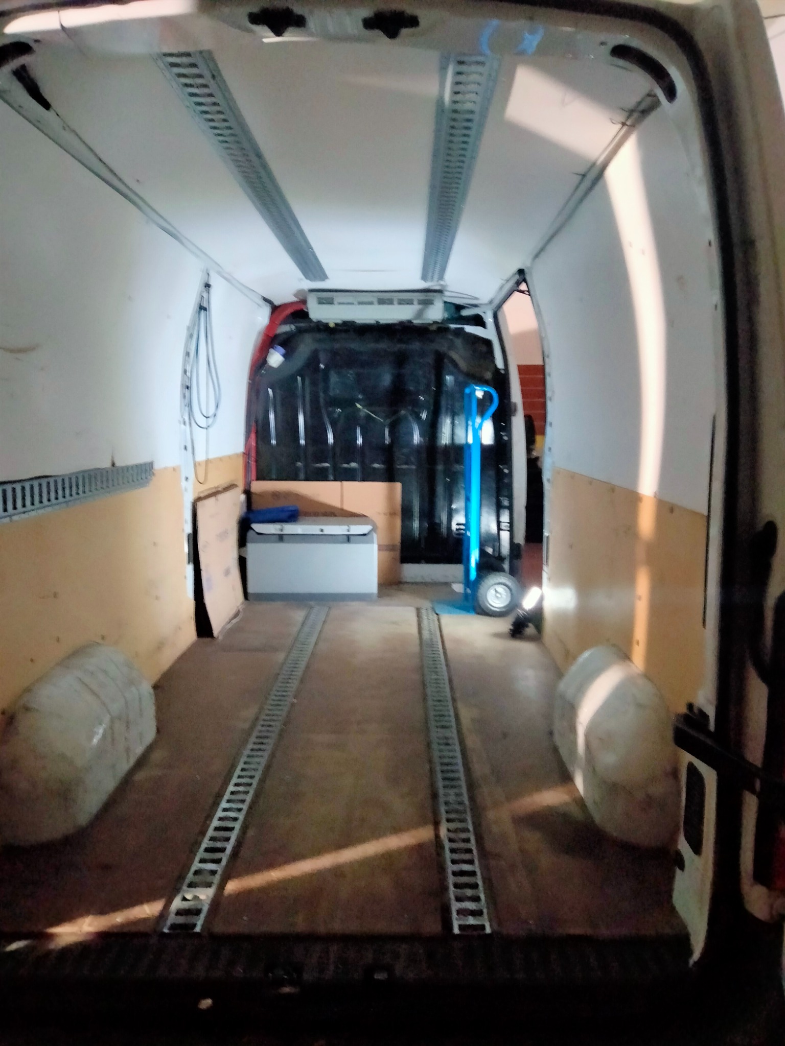Wnętrze furgonetki transportowej z zamontowanymi szynami mocującymi na podłodze i ścianach, widoczny wózek transportowy i skrzynia na narzędzia.