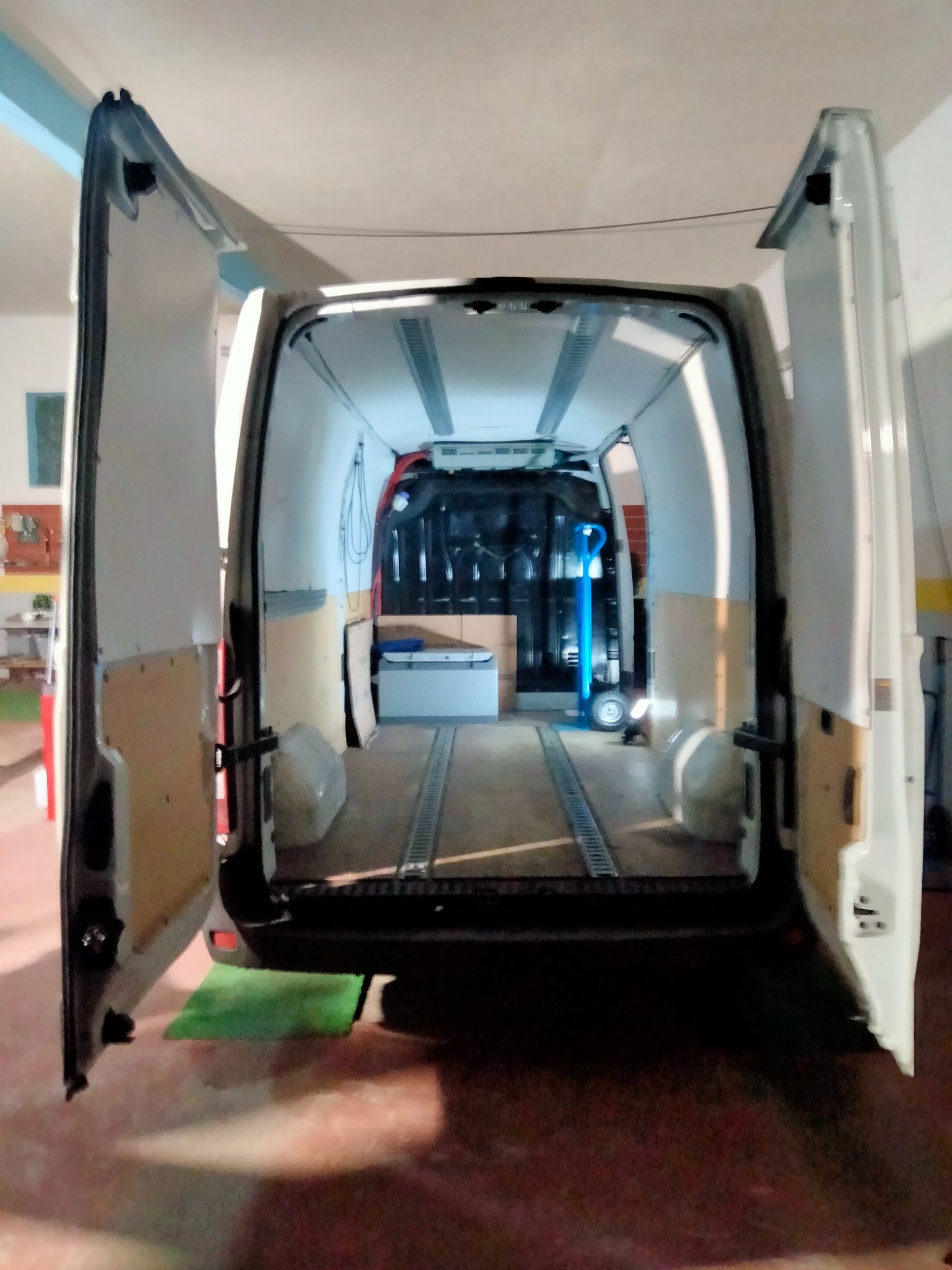 Wnętrze białego busa z otwartymi drzwiami, widoczne szyny podłogowe i wózek transportowy w środku, przygotowanego do załadunku.