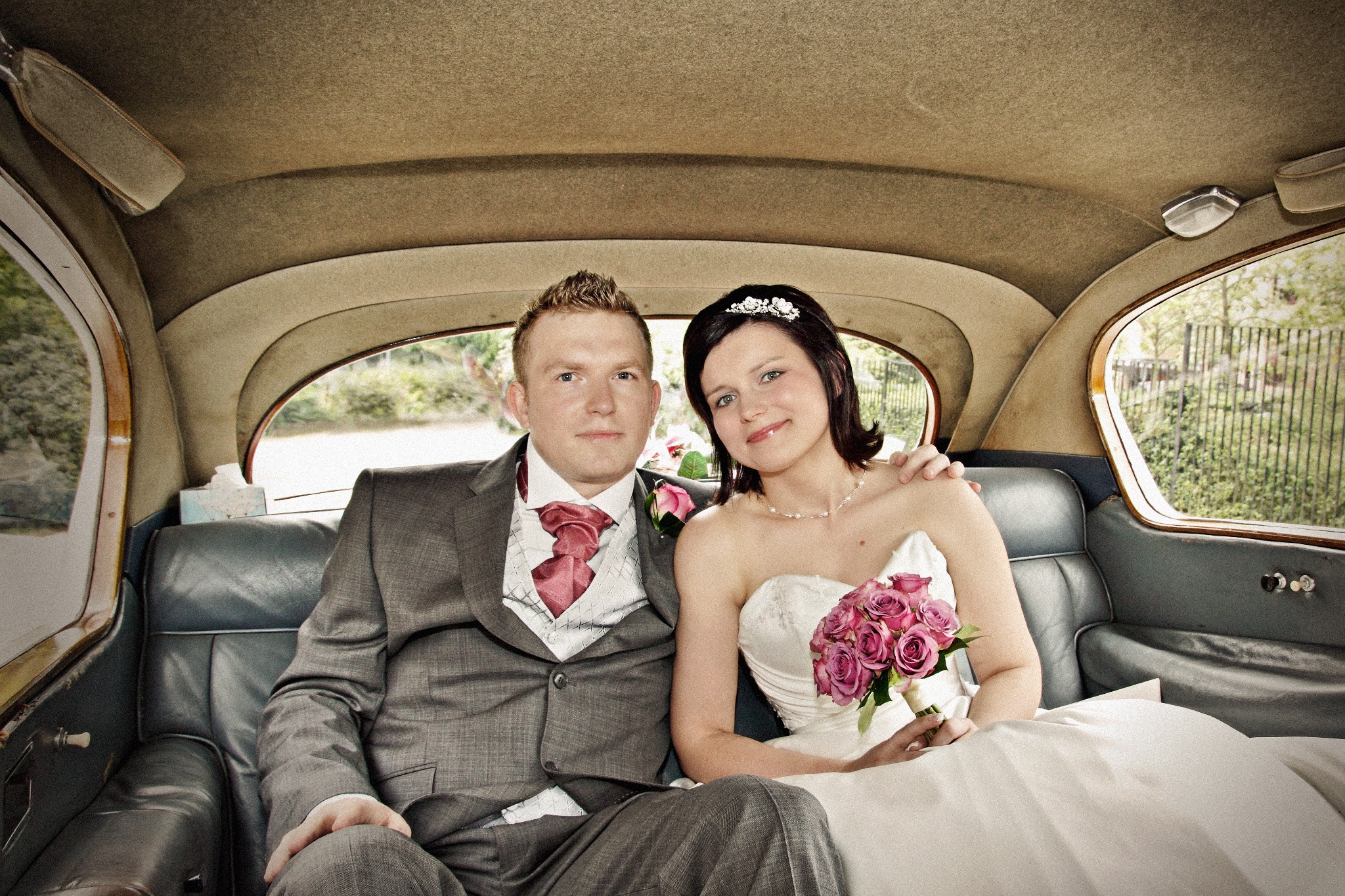 Para młoda w klasycznym samochodzie: pan młody w szarym garniturze z różową muszką i kamizelką, panna młoda w białej sukni bez ramiączek z bukietem różowych róż, siedząca na skórzanej kanapie...