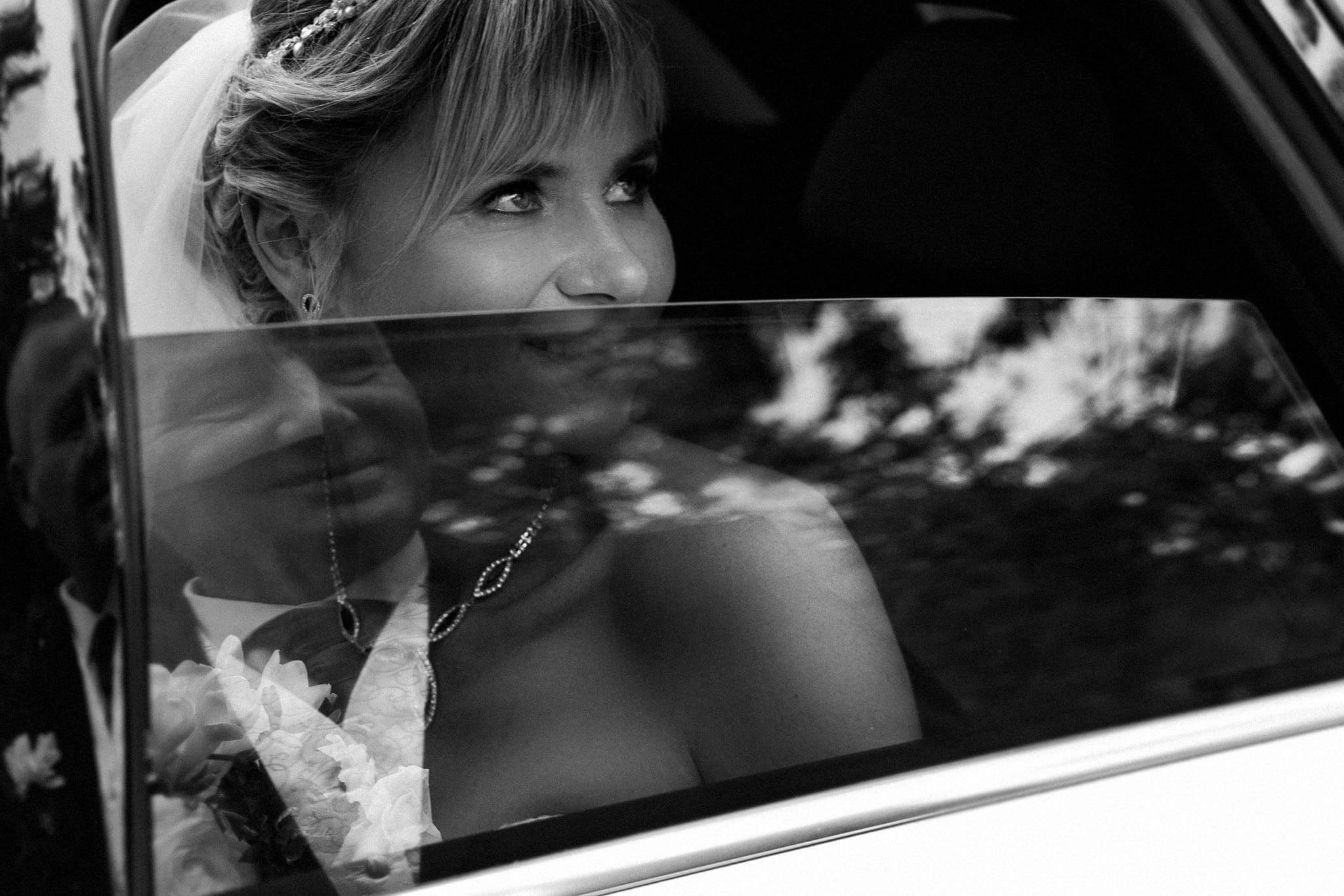 Czarno-białe zdjęcie panny młodej w welonie i z biżuterią, spoglądającej przez okno samochodu, z widocznym odbiciem pana młodego z bukietem.