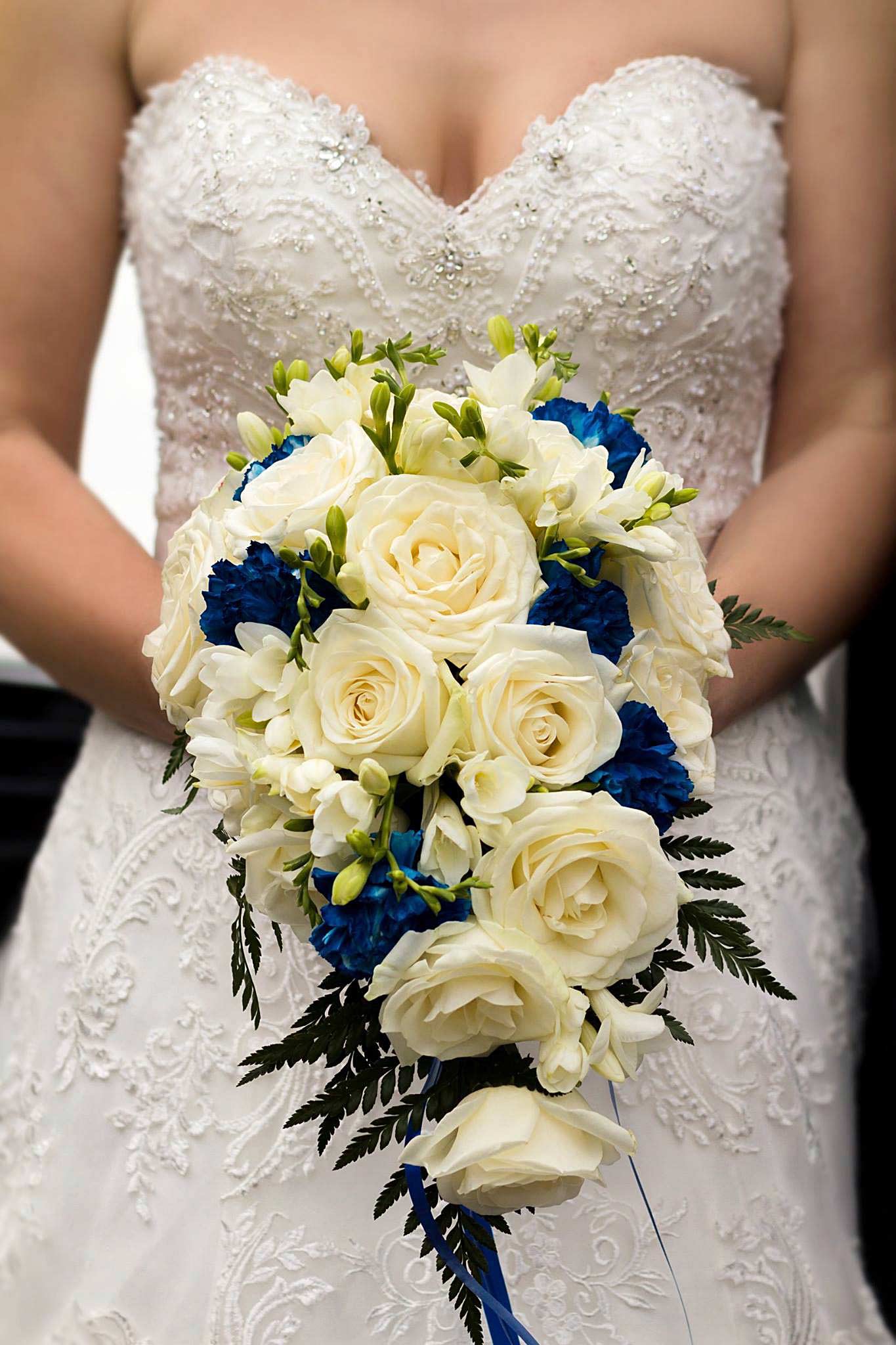 Panna młoda w koronkowej sukni trzyma bukiet ślubny z białych róż i niebieskich goździków.