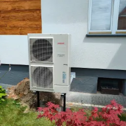 Biała pompa ciepła Atlantic Inverter zamontowana na czarnych wspornikach przy ścianie budynku z drewnianą okładziną i oknem, widoczny fragment trawnika i czerwony klon japoński.