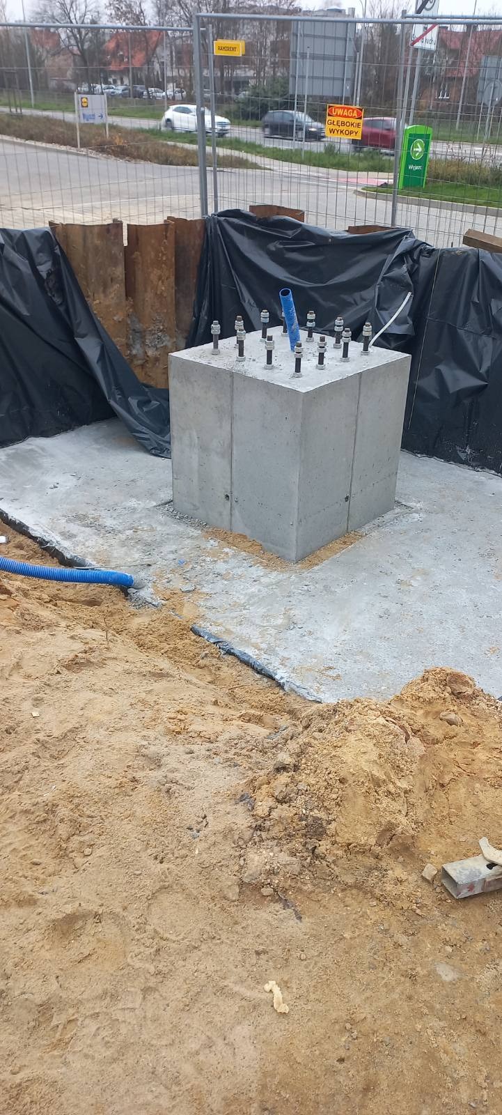 Betonowy blok fundamentowy z wystającymi śrubami kotwiącymi, otoczony wykopem zabezpieczonym folią i stalowymi szynami, widoczne rury instalacyjne.