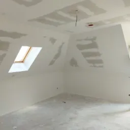 Poddasze po szpachlowaniu płyt kartonowo-gipsowych, widoczne szpachlowane łączenia na skosach i suficie, okno dachowe w ścianie, surowa podłoga przed dalszym wykończeniem.