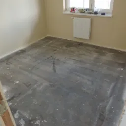 Przygotowane podłoże pod panele lub parkiet w pokoju z oknem, widoczne ślady po wcześniejszych pracach, ściany pomalowane na beżowo, na parapecie okiennym narzędzia i materiały budowlane.