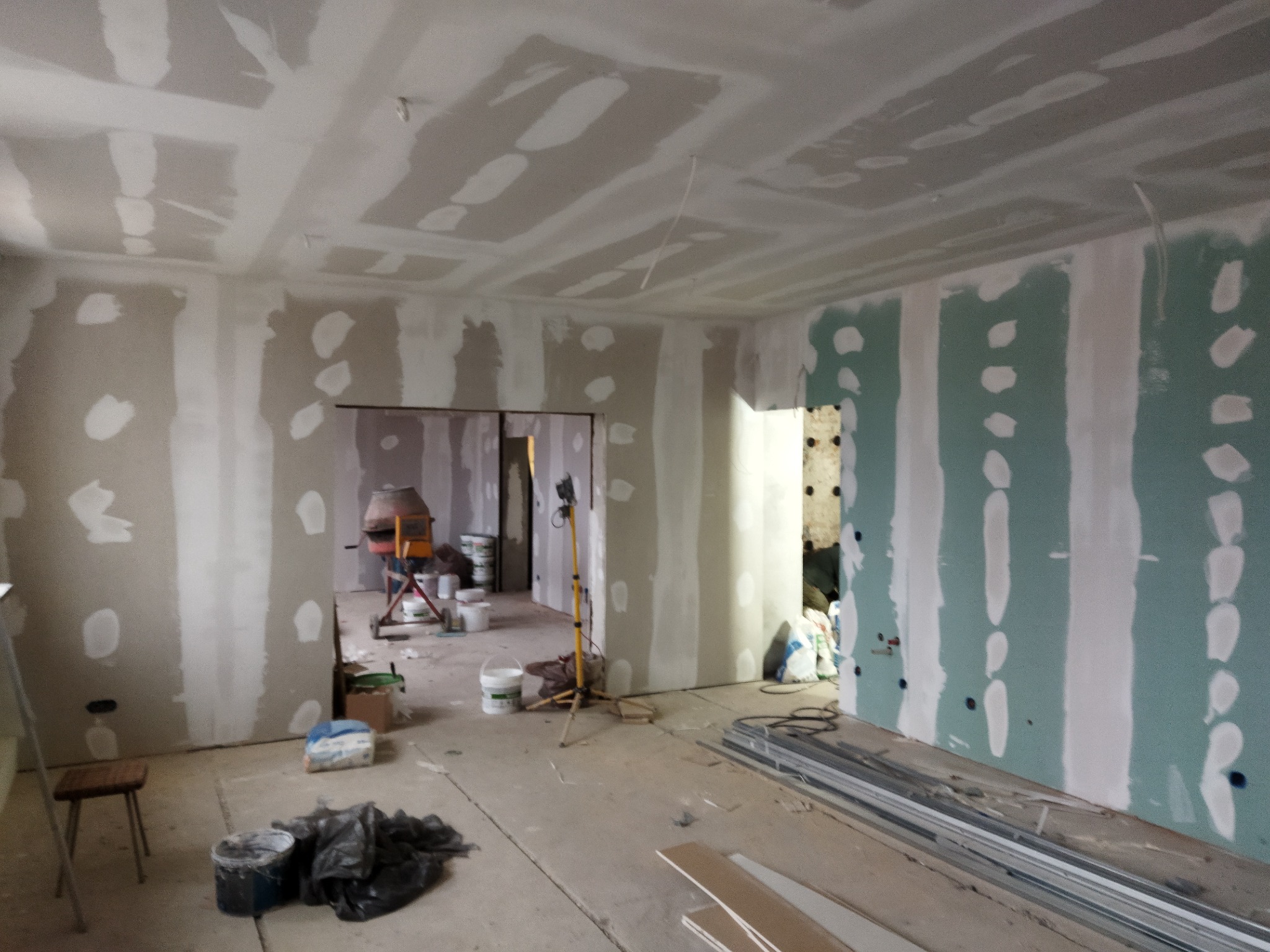 Wnętrze w trakcie prac wykończeniowych: ściany obłożone płytami gipsowo-kartonowymi, widoczne szpachlowanie łączeń, na podłodze leżą profile metalowe, w tle betoniarka i wiadra z zaprawą.