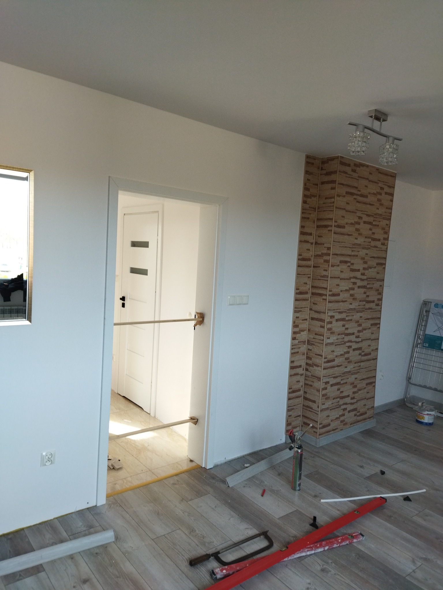 Wnętrze w trakcie remontu: widoczne narzędzia, poziomice, piła, listwy przypodłogowe, wykończone panele podłogowe, zabezpieczone wejście do pokoju oraz fragment dekoracyjnej ściany z imitacji cegły.