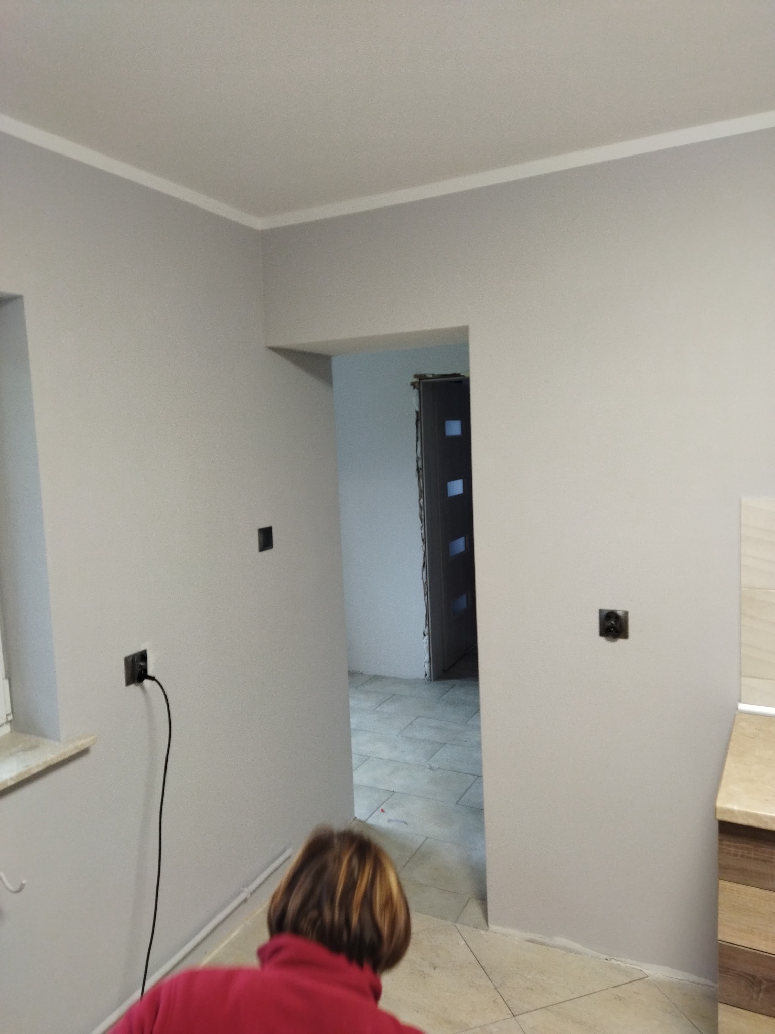 Świeżo pomalowane szare ściany z listwą przysufitową, widoczne gniazdka elektryczne i fragment osoby w czerwonej bluzie, podłoga wyłożona płytkami.